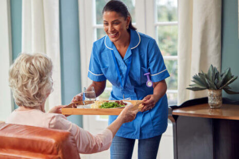Female Caregiver Bringing Meal On Tray To Senior Woman