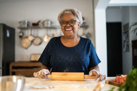 An older woman smiles as she rolls out pie crust, allowing her caregiver to practice self-care during Alzheimer’s by empowering her to handle tasks independently.