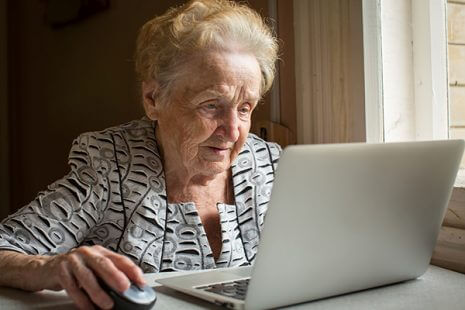 Elderly woman sitting at the table and types on laptop.