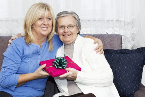 Smiling Mature nurse embracing senior woman and holding a Christmas gift while sitting on the sofa.