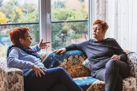 Two mature women are sitting on a sofa having a serious discussion about how to provide better senior care for an older parent.