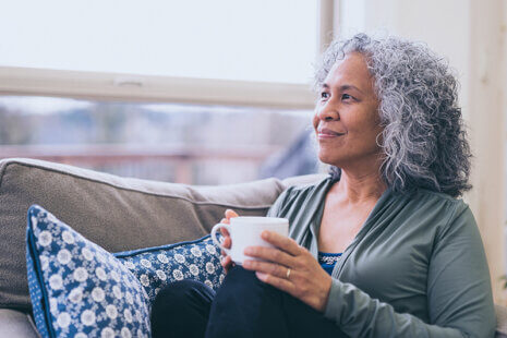 A mature woman who understands the importance of self-care for caregivers relaxes on a comfortable chair with pillows while holding a cup of coffee.