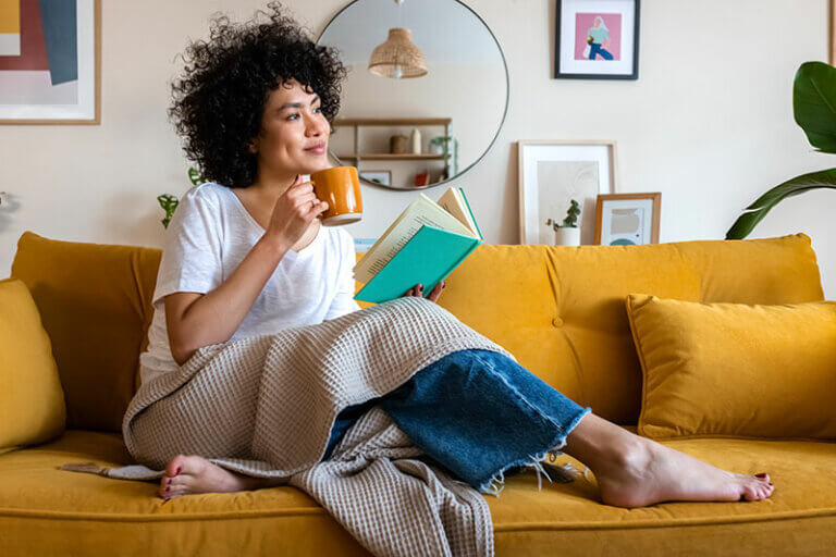 A woman sits comfortably on the couch, wrapped in a blanket and reading a book while holding a mug of coffee, knowing it’s important to make time for yourself as a caregiver.