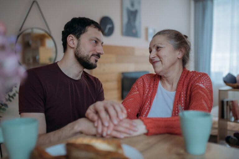 Adult son holding his mother's hand at the kitchen table, sharing a quiet moment during dementia care at home.